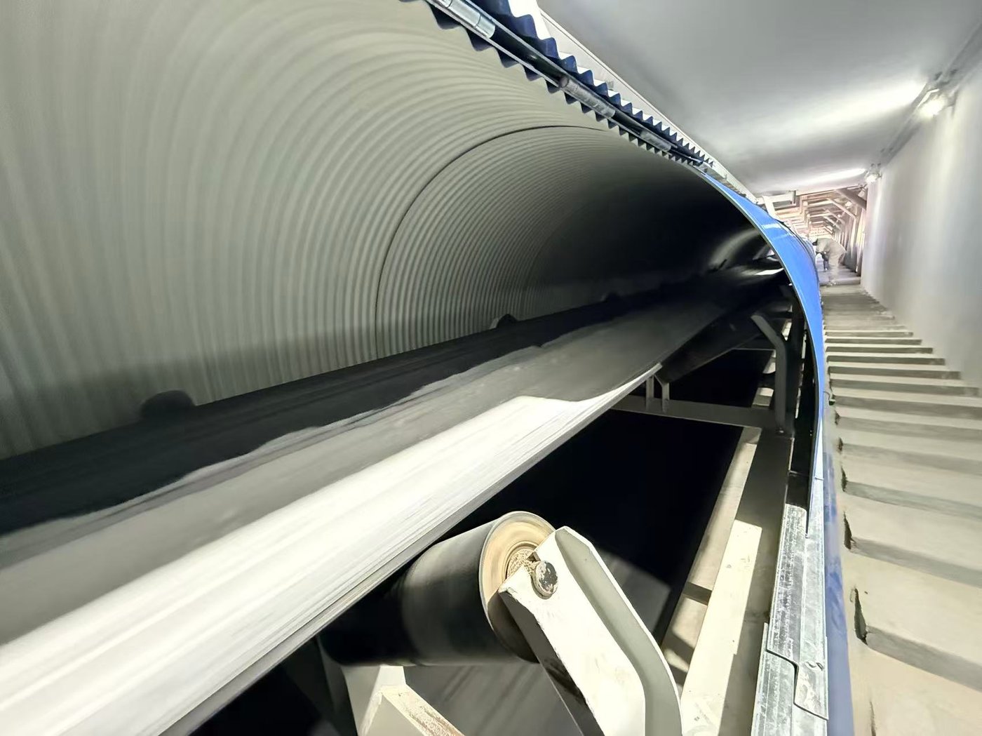 Interior of an enclosed belt conveyor gallery showing the troughed belt, carrying idlers and the blue cladding of the gallery
